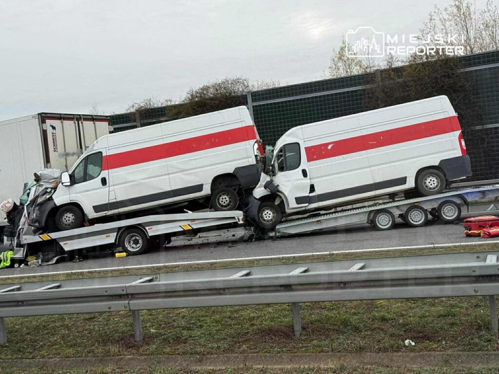 Dwa białe samochody dostawcze przewrócone na przyczepie transportowej na drodze, w otoczeniu barier ochronnych.