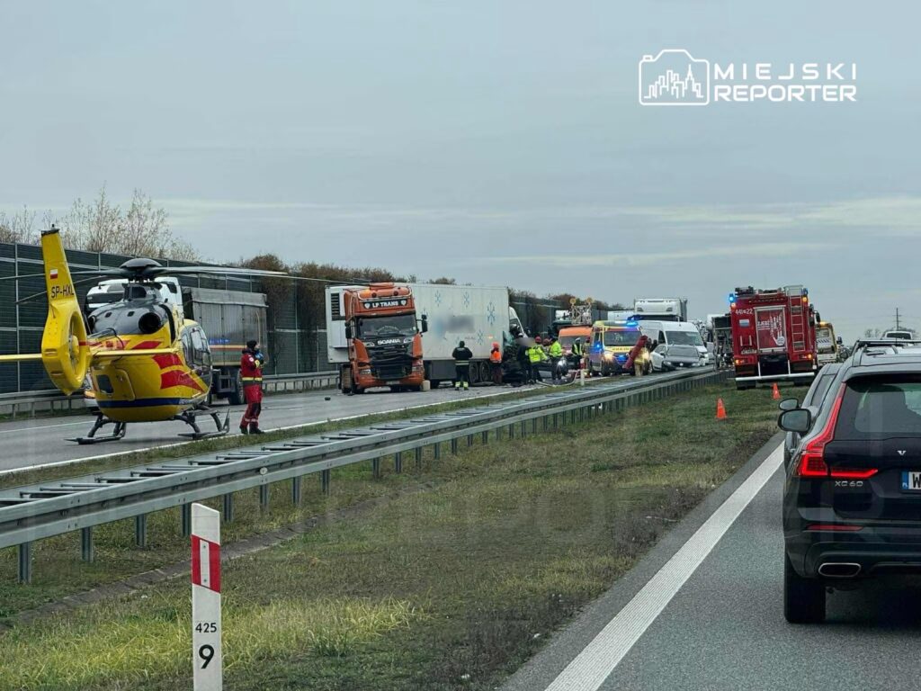Na drodze krajowej lądował helikopter ratunkowy, obok stoją pojazdy ciężarowe oraz samochody osobowe, a w tle widać służby ratunkowe.
