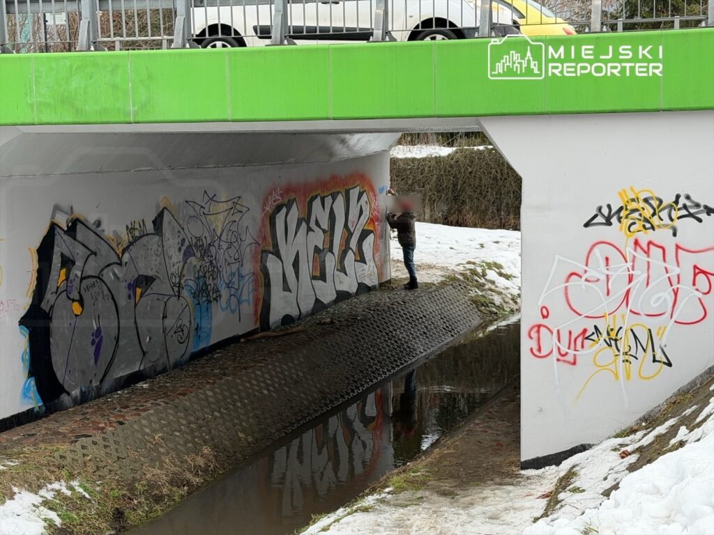 Mężczyzna stojący na brzegu ciekłej wody pod mostem, otoczony graffiti na ścianach i resztkami śniegu.
