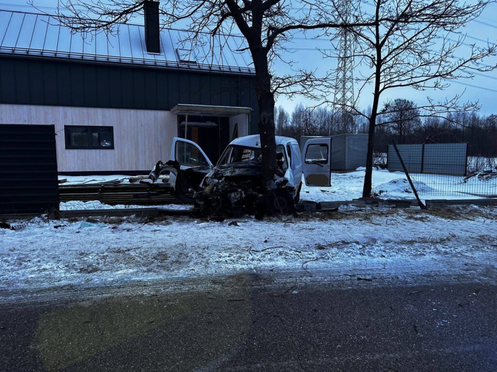 Rozbity samochód dostawczy uderzony w drzewo obok nowoczesnego budynku, otoczenie pokryte śniegiem.