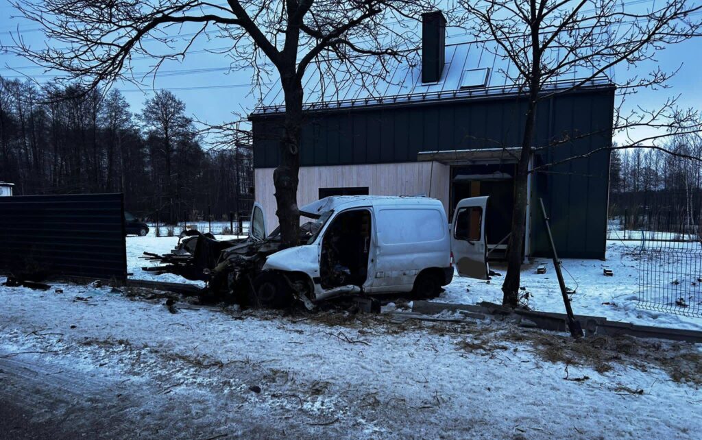 Zniszczony biały samochód dostawczy stoi obok drzewa, a w tle widoczny jest nowoczesny budynek w zimowym otoczeniu.