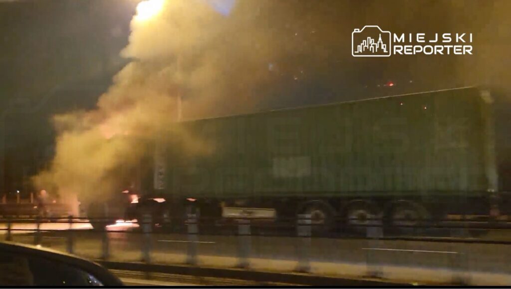 Zielona ciężarówka z kontenerem płonie na poboczu drogi, w otoczeniu gęstego dymu i jasnych płomieni.