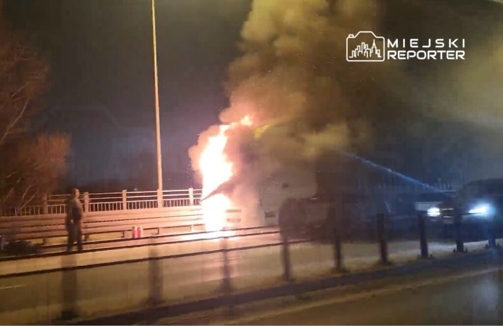 Pojazd płonący na poboczu ruchliwej drogi, otoczony gęstym dymem, w nocy.