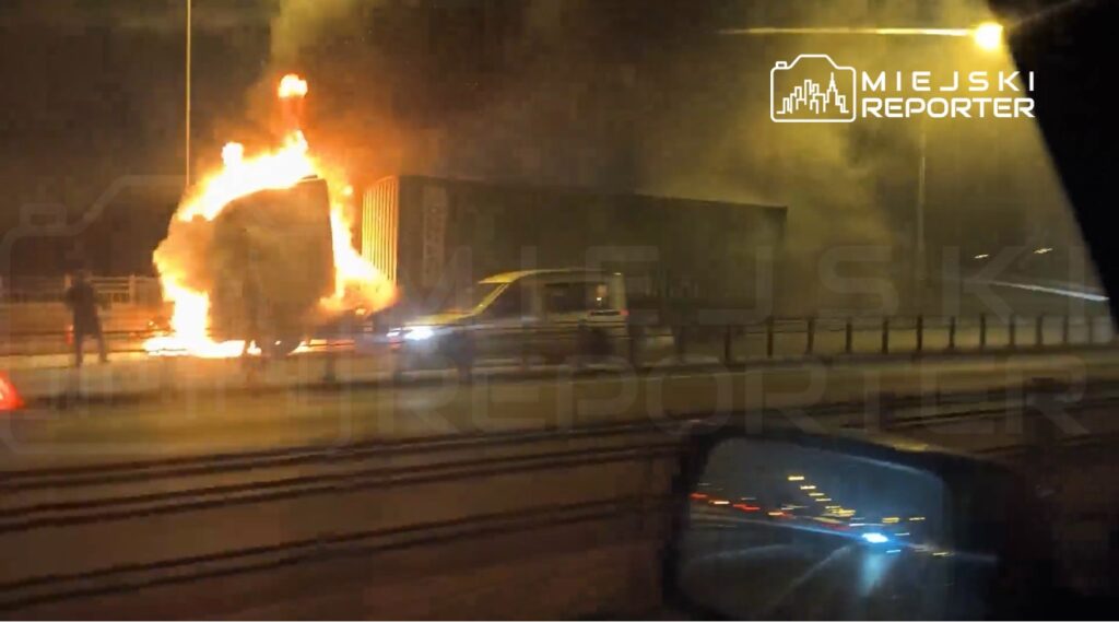 Pojazd ciężarowy płonący na drodze, obok stojący mężczyzna obserwujący zdarzenie w nocy.