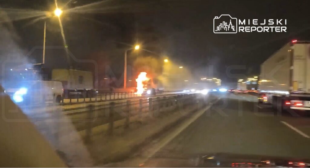 Pojazd na autostradzie płonie, wydobywają się z niego płomienie i dym, w tle widać inne samochody.