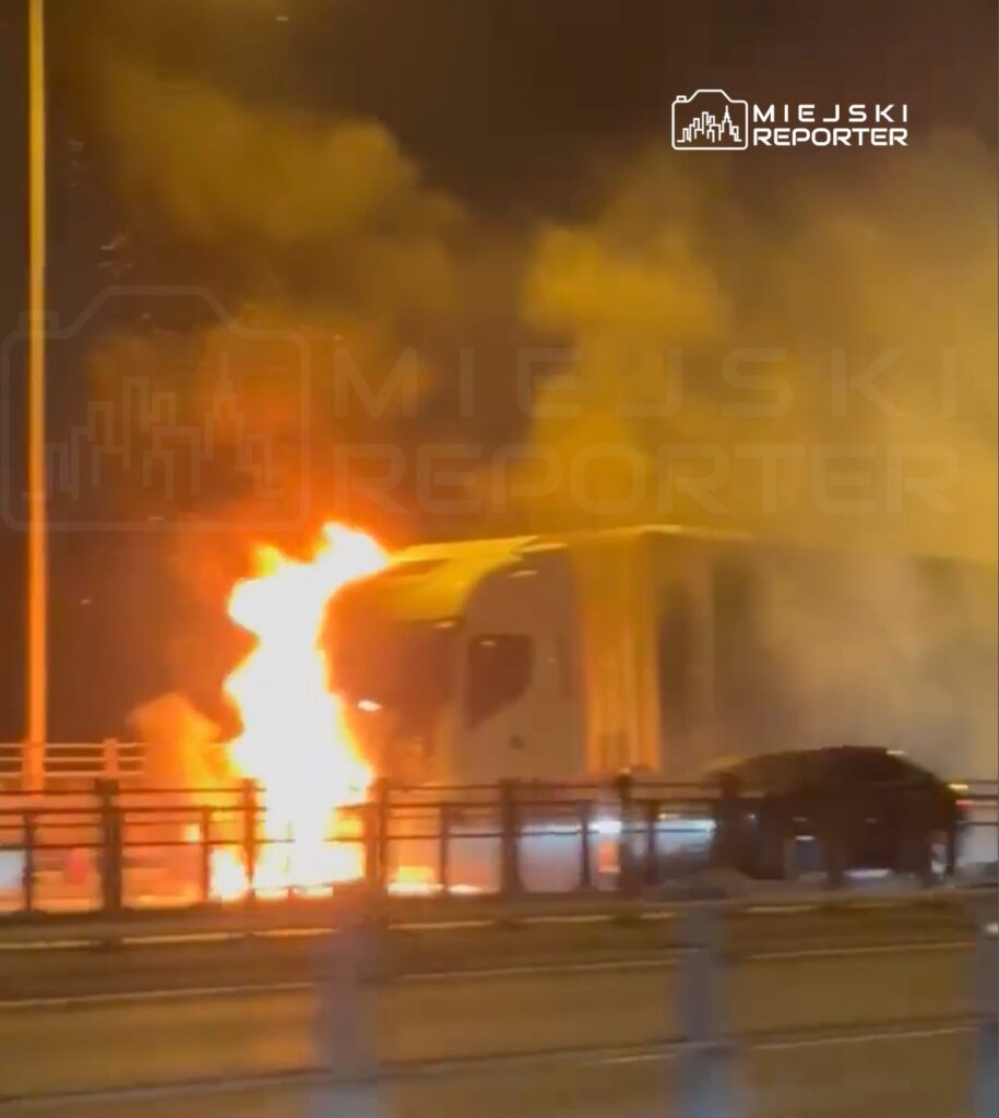 Ciężarówka płonąca na moście, otoczona gęstym dymem, obok przejeżdżający samochód.