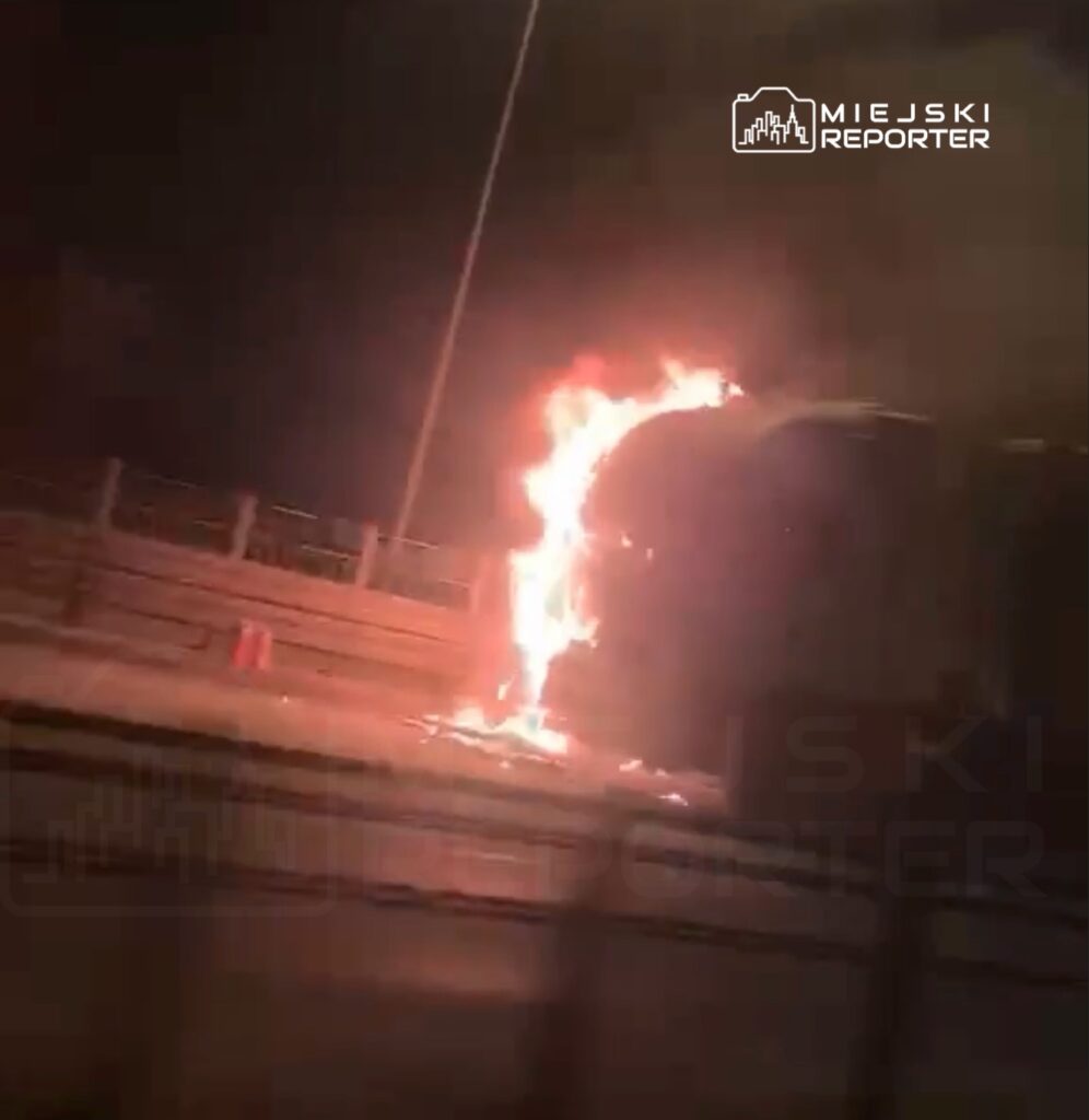 Pojazd płonący na poboczu drogi, otoczony dymem, w nocy. W tle widoczne bariery ochronne i światła uliczne.