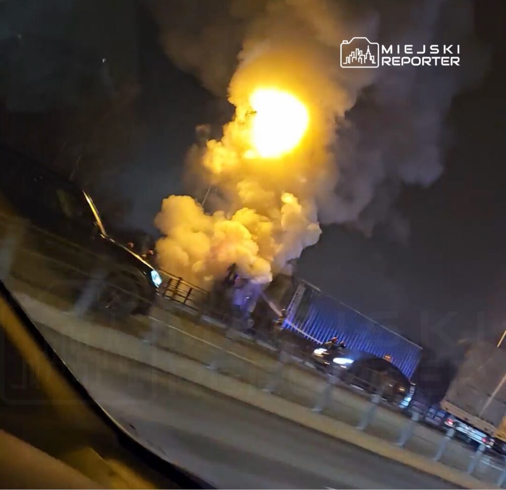 Wielka chmura dymu i ogień unoszą się nad jezdnią, obok stoją samochody w ruchu nocnym.