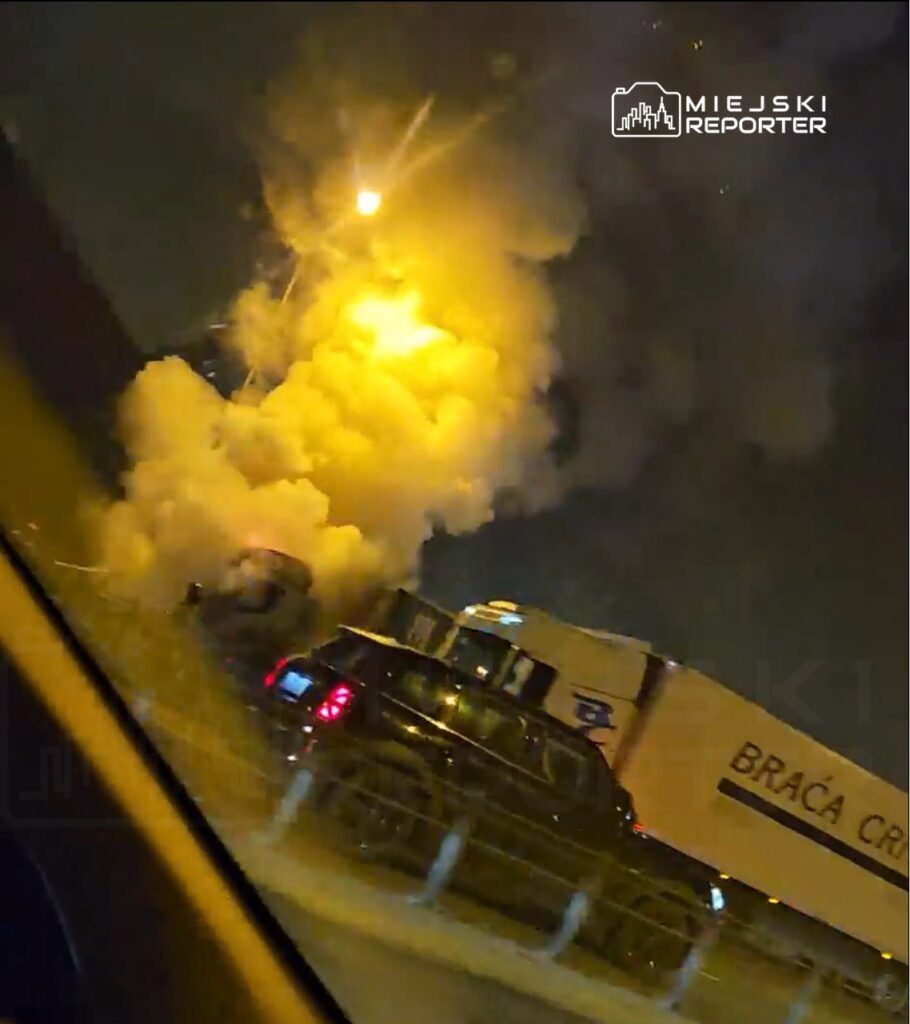 W nocy na drodze widać dym i ogień wydobywający się z pojazdu ciężarowego, otoczonego przez zaparkowane samochody.