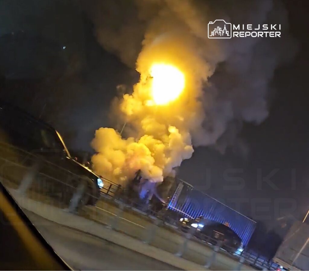 Wybuch gazu powodujący dużą chmurę dymu i ognia na drodze, w pobliżu zaparkowanych samochodów w nocy.