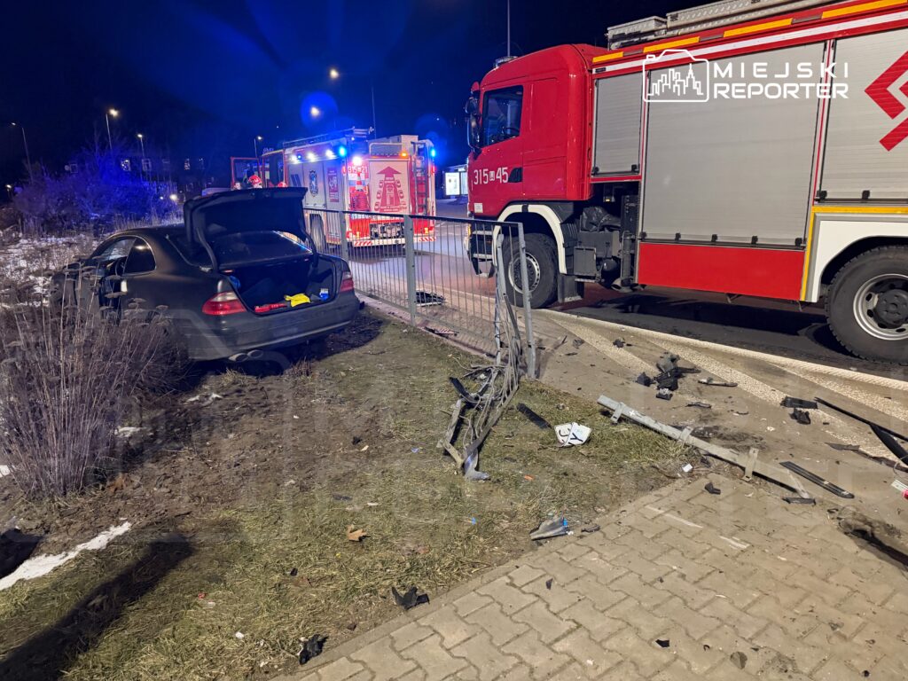 Czarny samochód osobowy z otwartą klapą bagażnika stoi na trawie obok uszkodzonego ogrodzenia, w pobliżu wozu strażackiego.