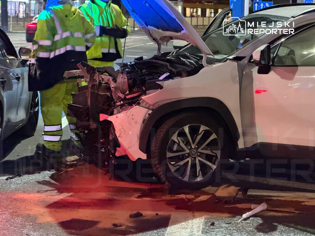 Dwaj mężczyźni w odblaskowych kurtkach badają uszkodzony przód białego SUV-a na ulicy nocą.