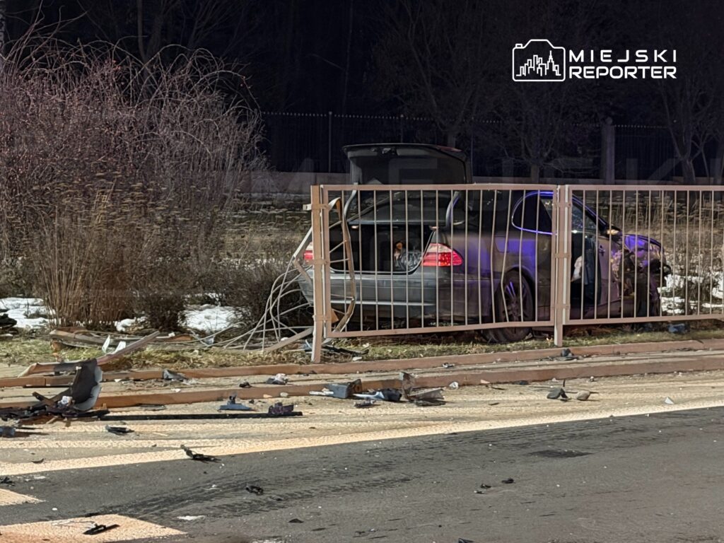 Uszkodzony samochód osobowy stoi za ogrodzeniem, otwarta klapa bagażnika, a wokół leżą fragmenty wraku.