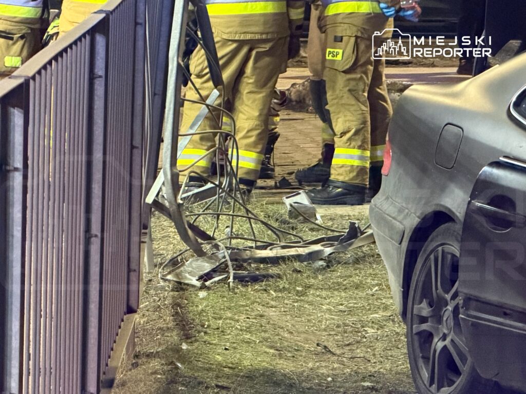 Grupa strażaków w mundurach z odblaskowymi elementami pracuje przy zniszczonym ogrodzeniu, obok uszkodzonego samochodu.