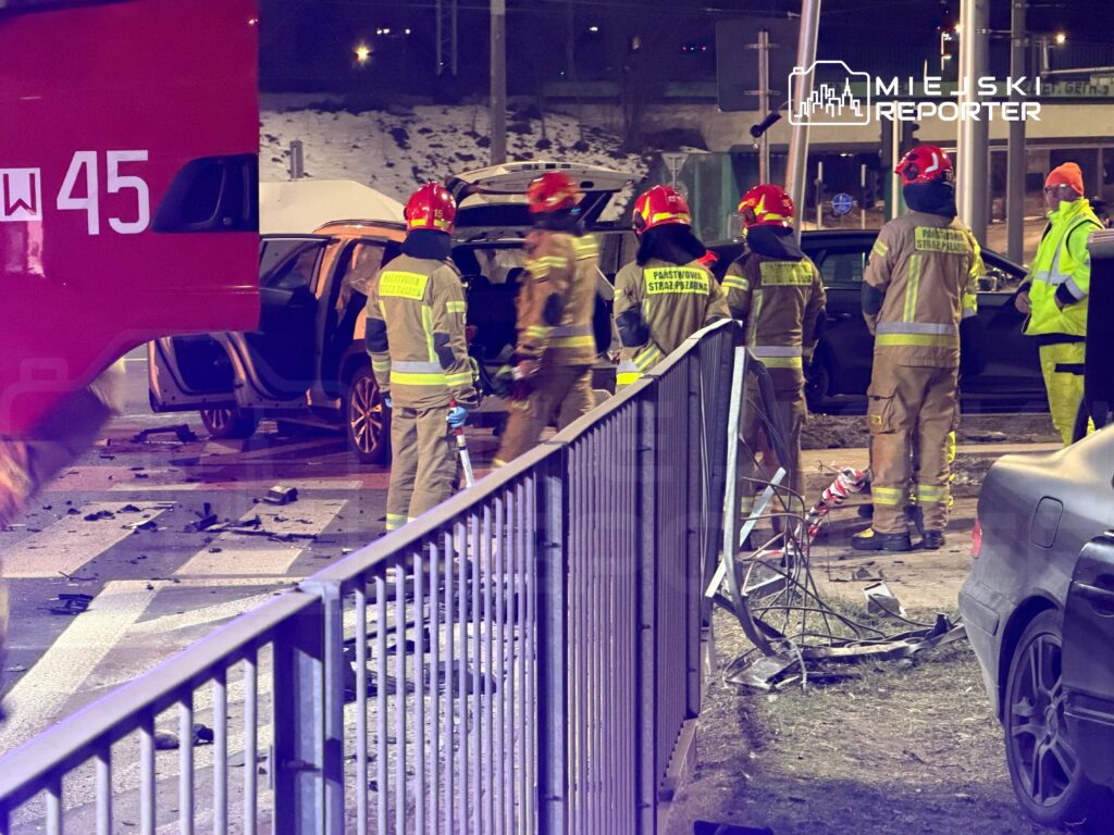 Grupa strażaków w odblaskowych strojach bada miejsce wypadku samochodowego na ruchliwej ulicy. W tle uszkodzone pojazdy.