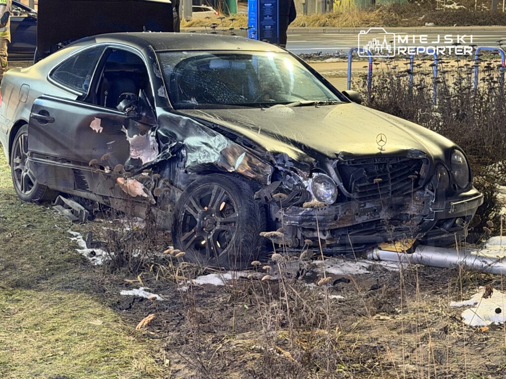 Zniszczony czarny samochód marki Mercedes leży na poboczu drogi, obok widoczne uszkodzenia i roślinność.