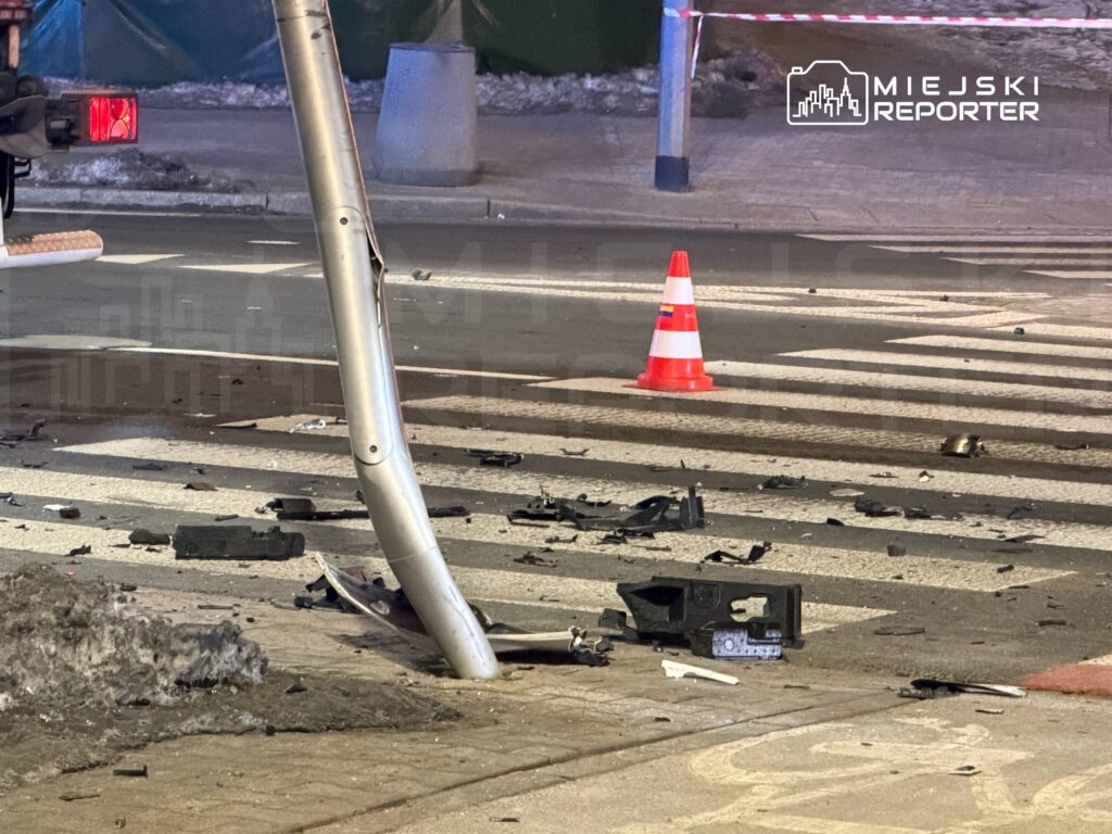 Na jezdni leżą resztki pojazdu, a obok uszkodzony słup i pachołek ostrzegawczy.