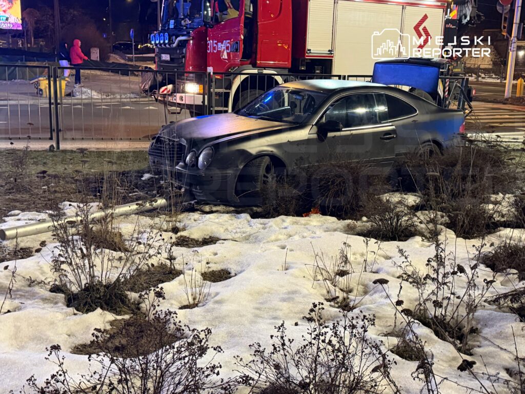 Czarny samochód osobowy zaparkowany w zaroślach na skraju jezdni, obok wozu strażackiego. W tle widoczni przechodnie.