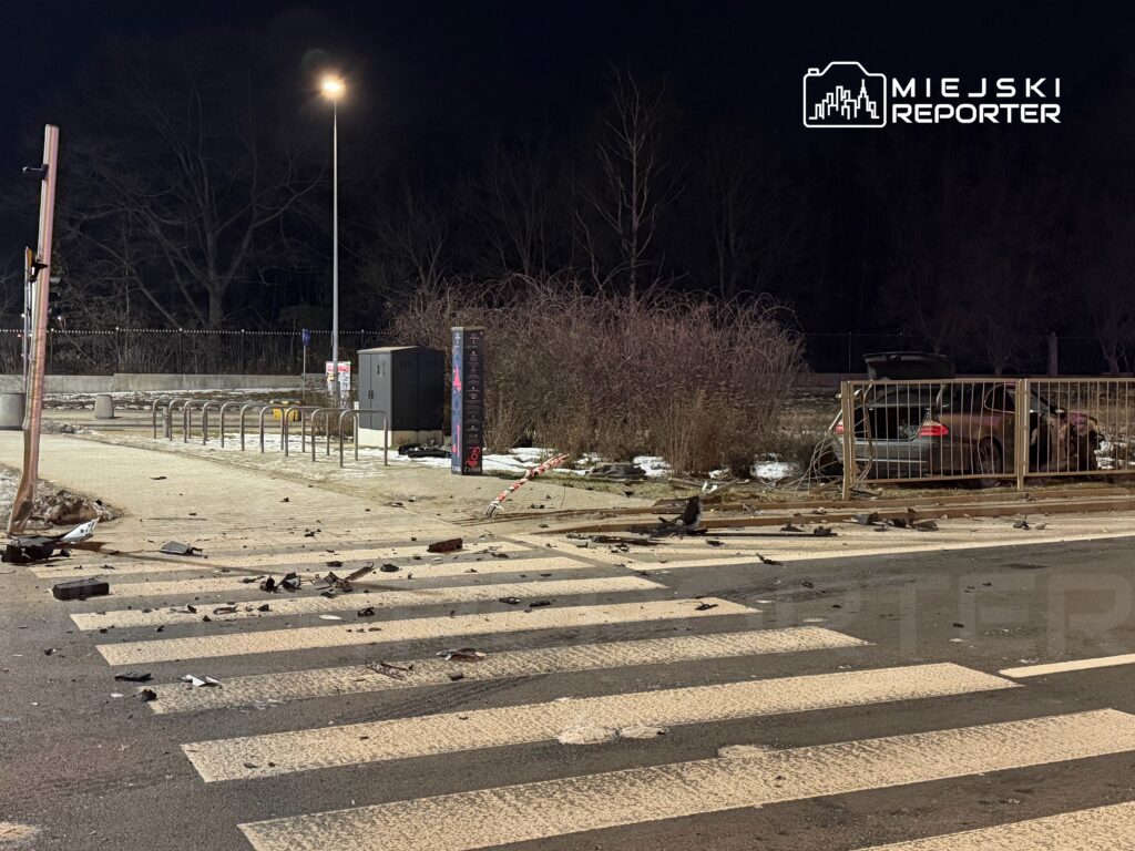 Zniszczona nawierzchnia drogi z fragmentami pojazdów i barierkami, obok zaparkowane auto w ciemnym otoczeniu.