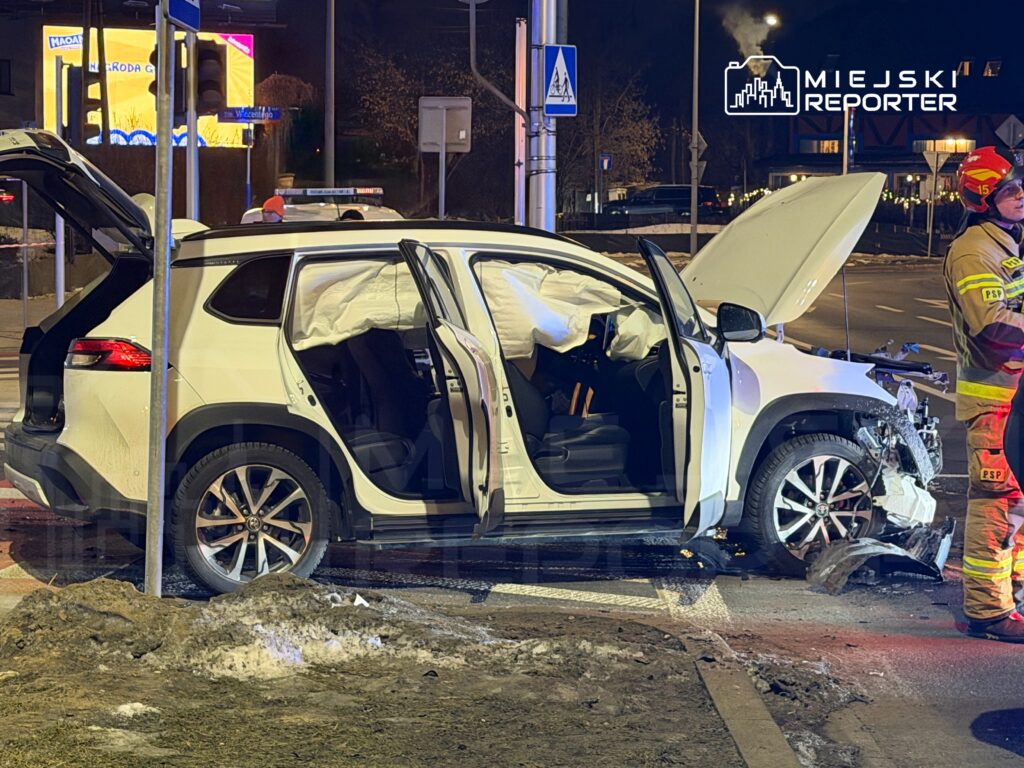 Biały samochód osobowy z otwartymi drzwiami stoi na poboczu drogi, a obok niego strażak w mundurze interweniuje po wypadku.