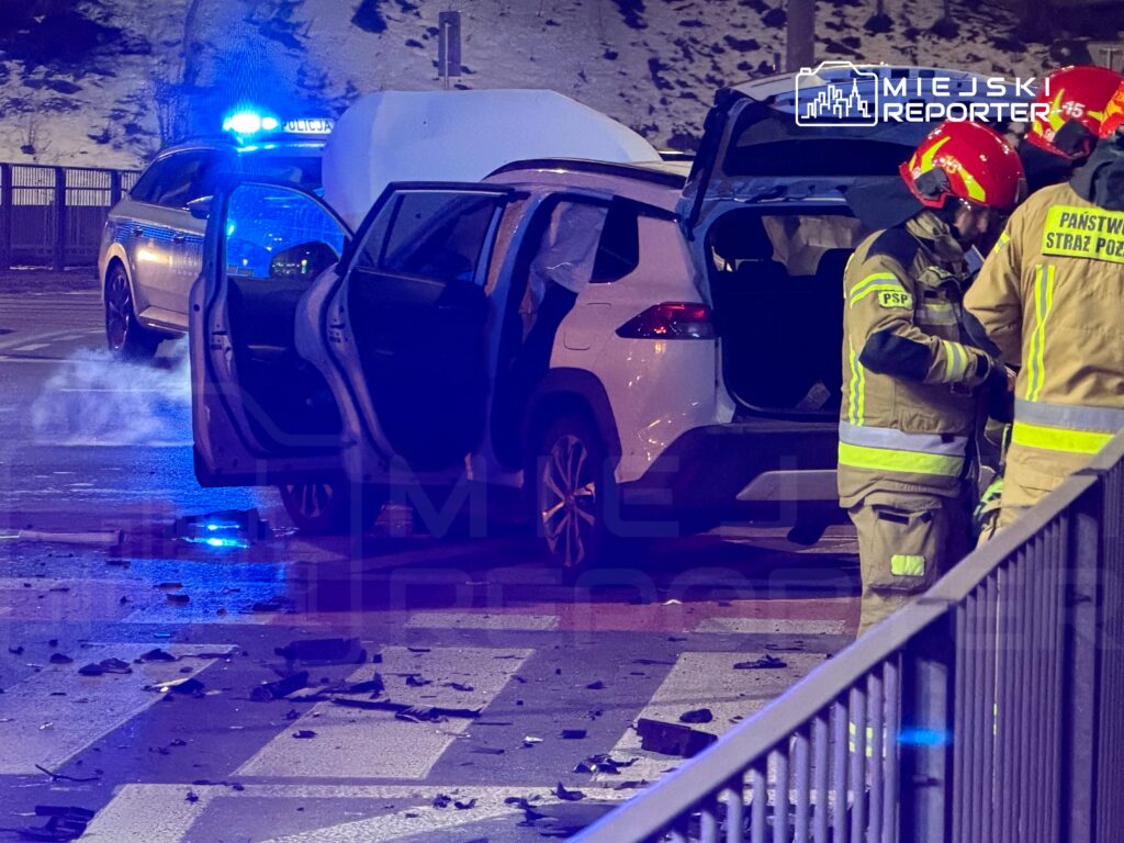 W nocy, w tle niebieskie światła policyjne, strażacy pracują przy rozbitej samochodowej na jezdni, obok leżą fragmenty pojazdu.