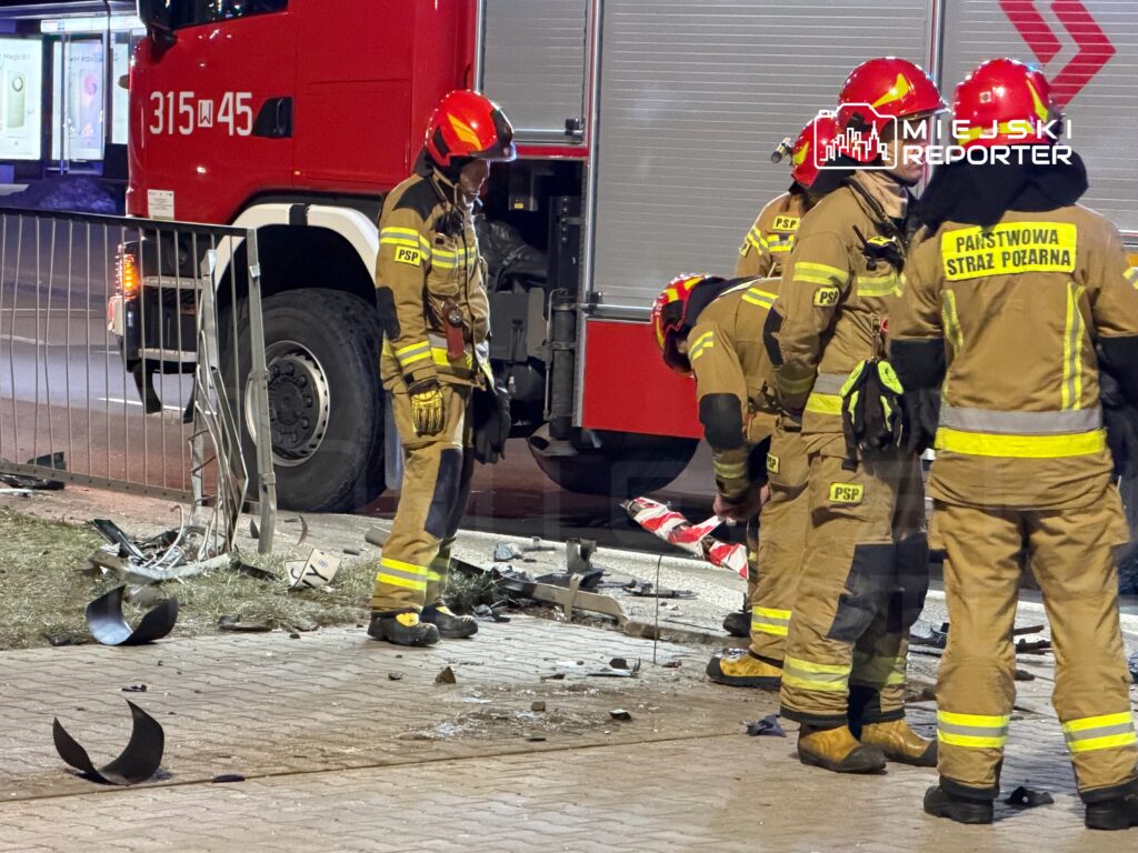 Grupa strażaków w mundurach pracuje przy usuwaniu skutków wypadku na jezdni, obok wozu strażackiego.