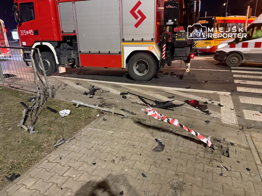 Uszkodzony fragment drogi z rozrzuconymi odłamkami oraz czerwonym wozem strażackim zaparkowanym obok.