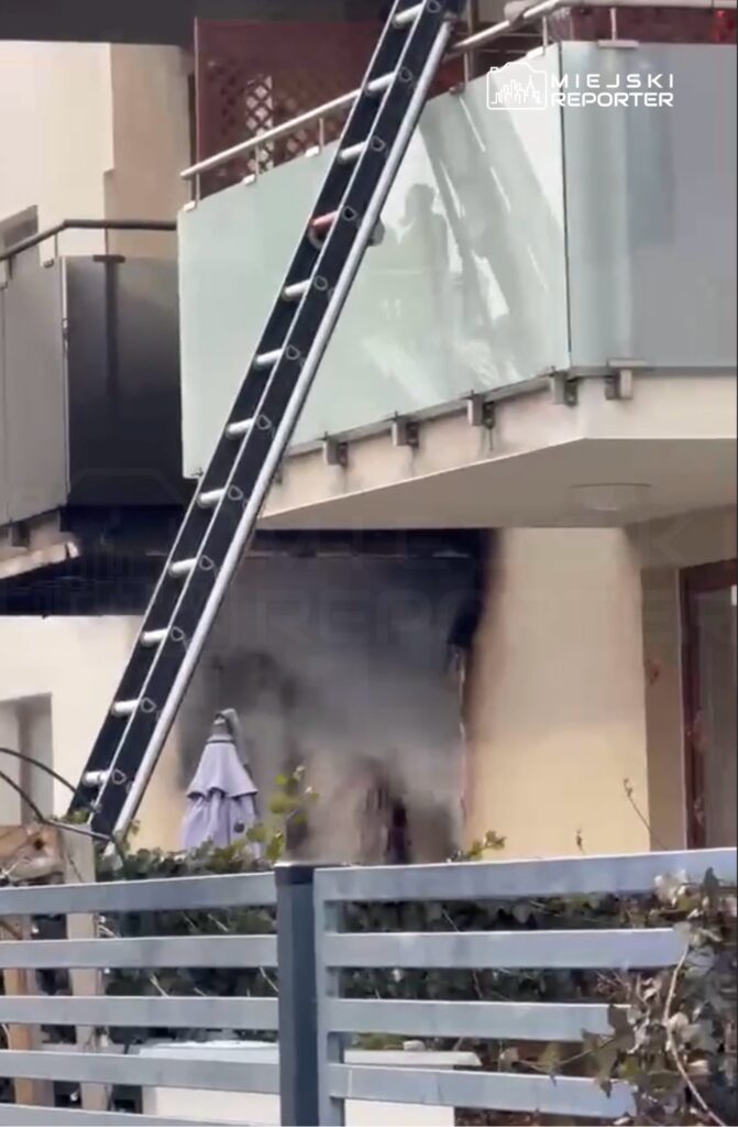 Dym wydobywający się z balkonu mieszkania, obok stoi drabina przystawna, w tle parasol ogrodowy.