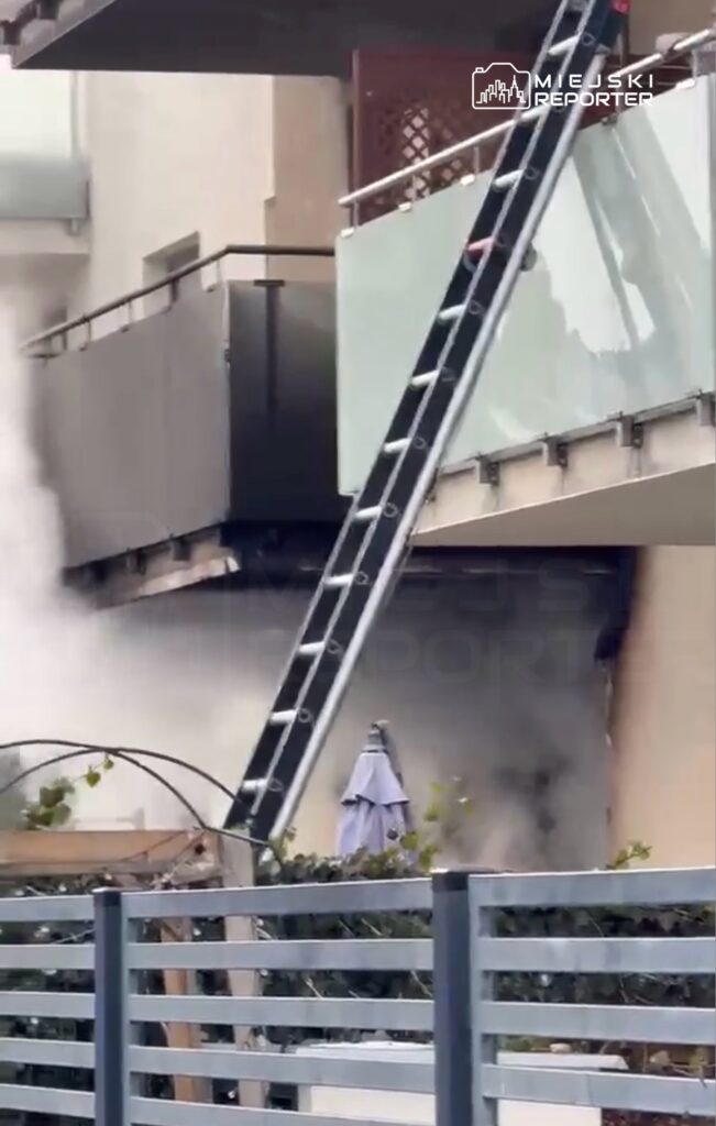 Dym wydobywający się z okna mieszkania, obok drabina oparta o balkon, w tle roślinność na ogrodzeniu.