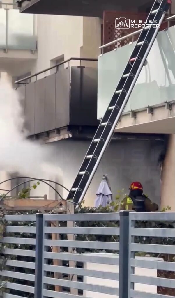 Strażak w hełmie używa drabiny, aby dotrzeć do miejsca pożaru na balkonie budynku, z którego wydobywa się dym.