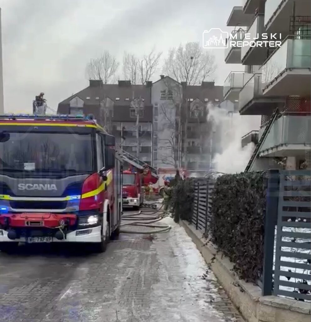Czerwony wóz strażacki z niebieskimi światłami stoi na ulicy, a w tle widać unoszący się dym z budynku mieszkalnego.