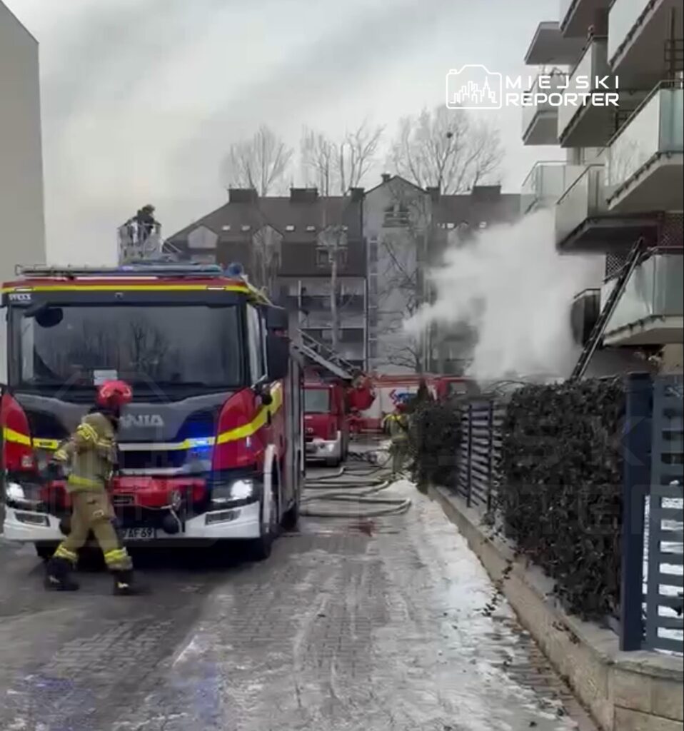 Strażak w hełmie i mundurze przechodzi obok wozu strażackiego na tle dymu wydobywającego się z budynku mieszkalnego.