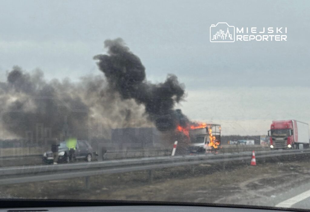 Pojazd osobowy zatrzymany na poboczu drogi, obok płonący ciężarówka wydobywająca czarny dym.