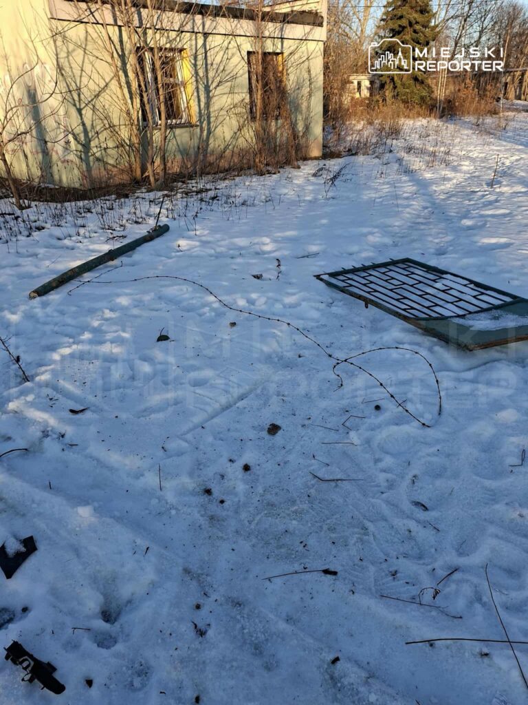 Zniszczony budynek z oknami bez szyb otoczony śniegiem i suchą roślinnością, z wystającymi elementami metalowymi.