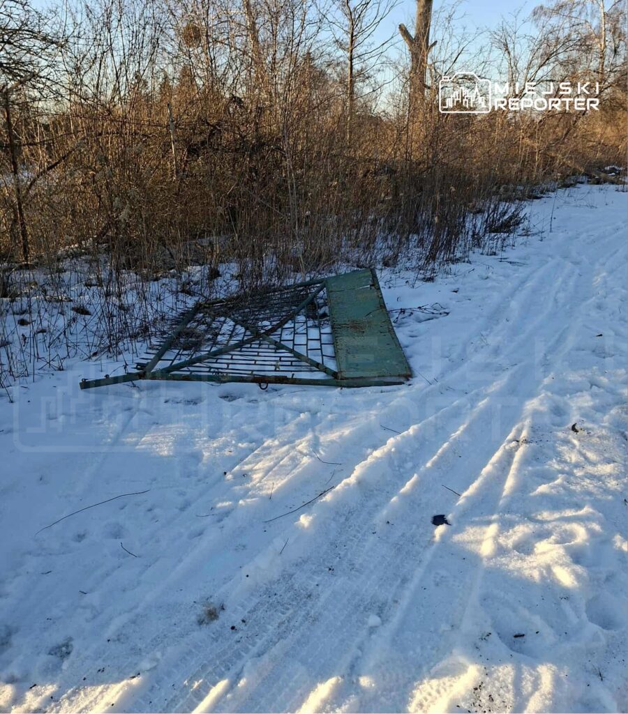 Zardzewiała brama leży na śniegu obok leśnej ścieżki, otoczona krzakami i drzewami.