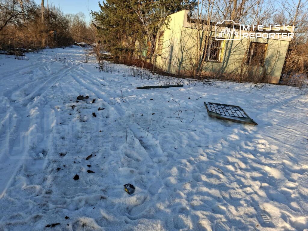 Zasypana śniegiem ścieżka prowadząca do opuszczonego, zielonego budynku otoczonego zaroślami.