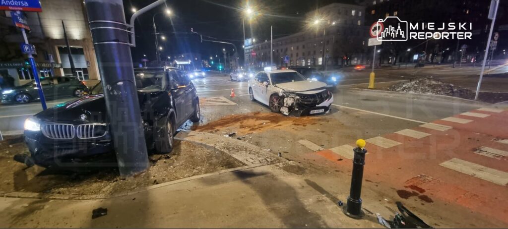 Dwa uszkodzone samochody osobowe, czarny SUV uderzony w latarnię, biały pojazd taksówki z rozbitym przodem na skrzyżowaniu.