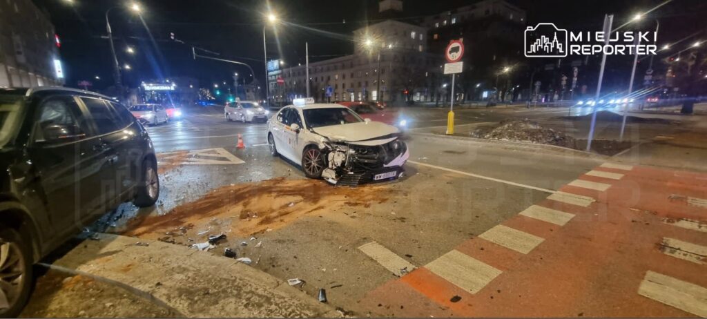 Wgnieciony przód białego samochodu osobowego na skrzyżowaniu, obok leży rozbity element nadwozia.