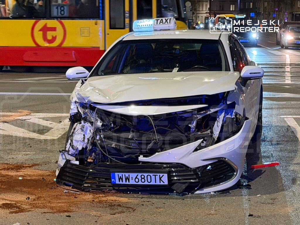 Rozbity samochód osobowy z uszkodzoną przednią częścią stoi na skrzyżowaniu, obok przystanku tramwajowego.