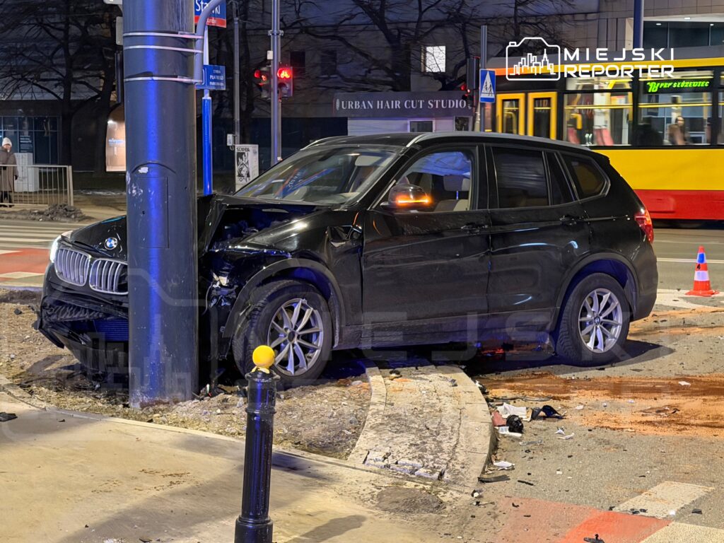Czarny SUV uderzył w słup oświetleniowy na skrzyżowaniu, obok widoczna jest zniszczona nawierzchnia i oznakowanie drogowe.