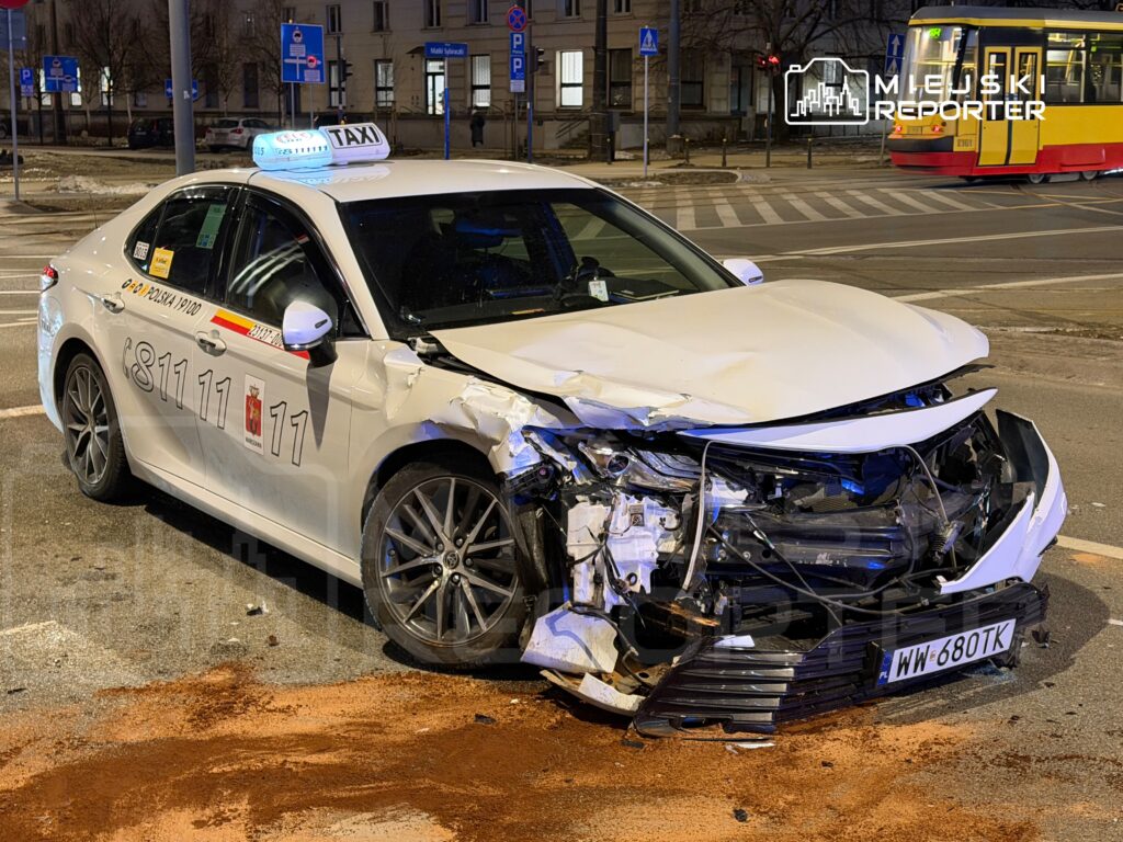 Uszkodzone taxi z wgniecionym przodem stoi na skrzyżowaniu, obok widoczny tramwaj oraz znaki drogowe.