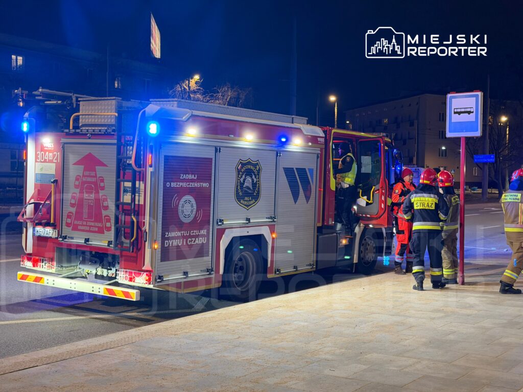 W nocy przy wozie strażackim z włączonymi sygnałami świetlnymi stoją strażacy w mundurach, rozmawiając o akcji ratunkowej.