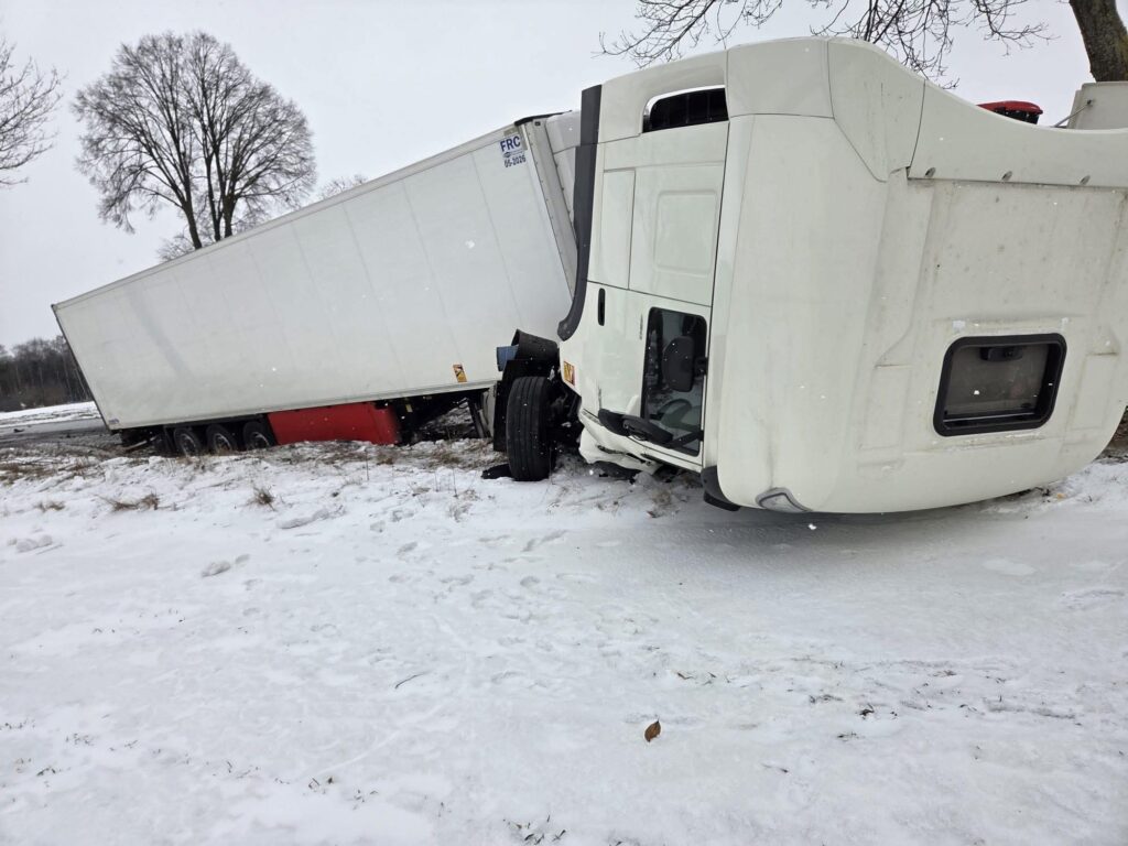 Ciągnik siodłowy leży na boku w zaśnieżonym terenie, z przyczepą częściowo w rowie. W tle widoczne drzewa.