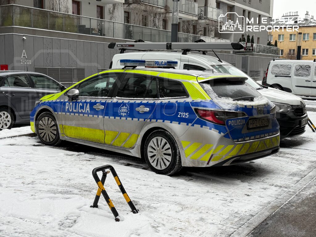 Policyjny samochód zaparkowany na zaśnieżonej ulicy obok innych pojazdów.