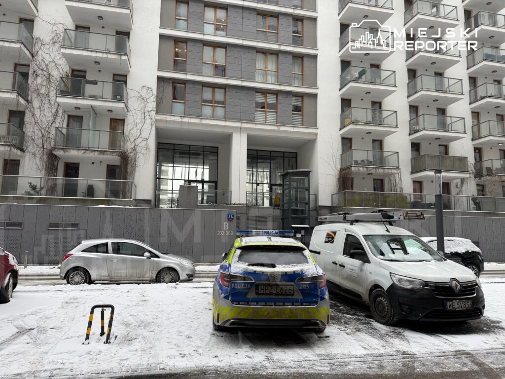 Policyjny radiowóz zaparkowany na zaśnieżonej ulicy przed nowoczesnym budynkiem mieszkalnym z przeszkloną fasadą.