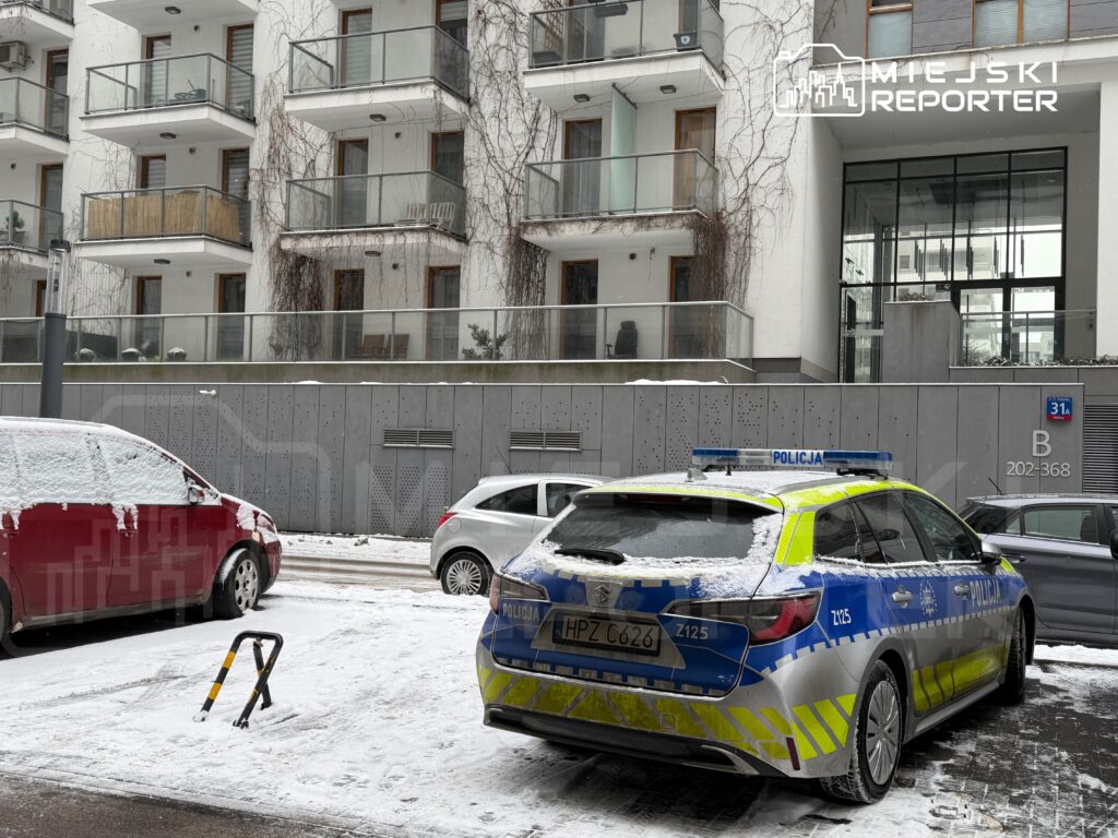 Policyjny radiowóz zaparkowany na zaśnieżonej ulicy obok czerwonego samochodu, w tle nowoczesny budynek mieszkalny.