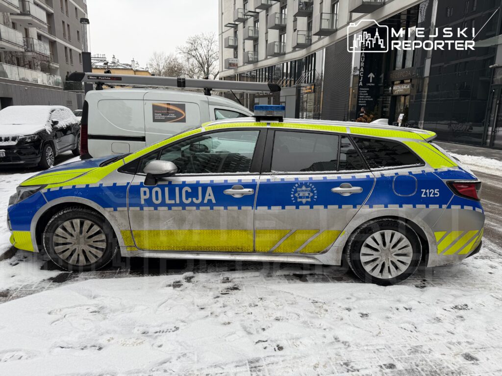 Radiowóz policyjny zaparkowany na zaśnieżonej ulicy w mieście, obok białego furgonu.