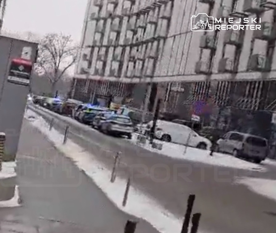 Policyjna akcja w mieście, z radiowozami zaparkowanymi wzdłuż ulicy obok nowoczesnego budynku.