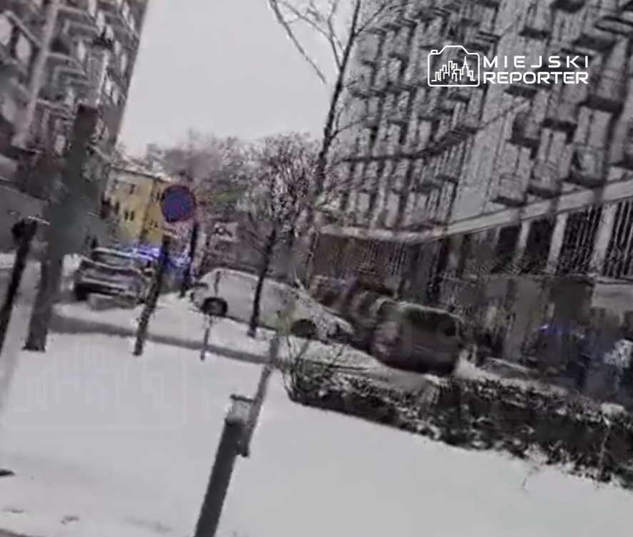 Zasypana śniegiem ulica z nowoczesnymi budynkami w tle, widać radiowóz z włączonymi sygnalizatorami.