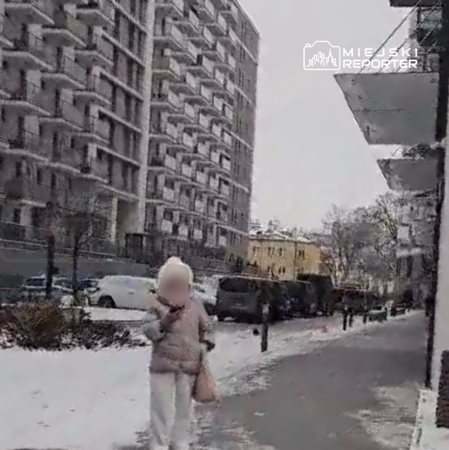 Kobieta w jasnej kurtce spacerująca po ośnieżonej ulicy, obok nowoczesnego budynku mieszkalnego.